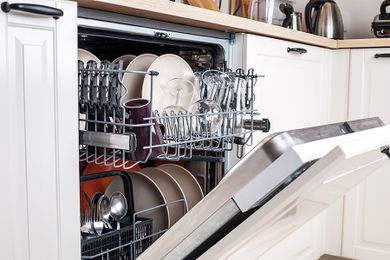 A dishwasher slightly ajar, filled with plates, bowls, cups, and silverware