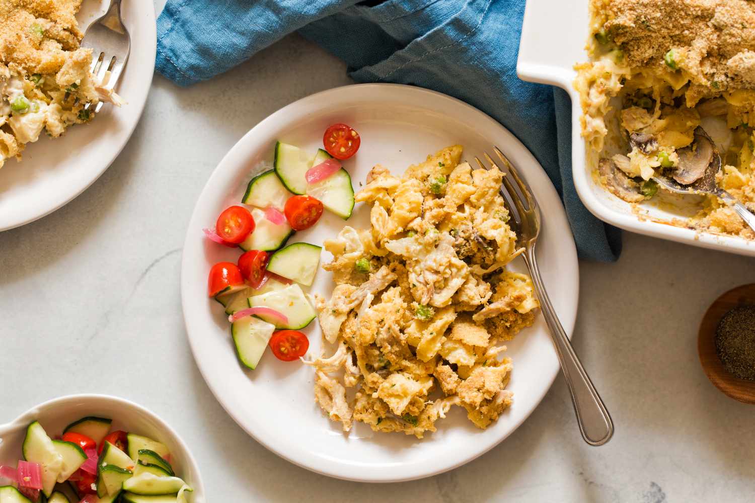 Overhead view of a white plate of chicken tetrazzini, cucumber and tomato salad, and fork next to a dish of tetrazzini and a blue cloth napkin 