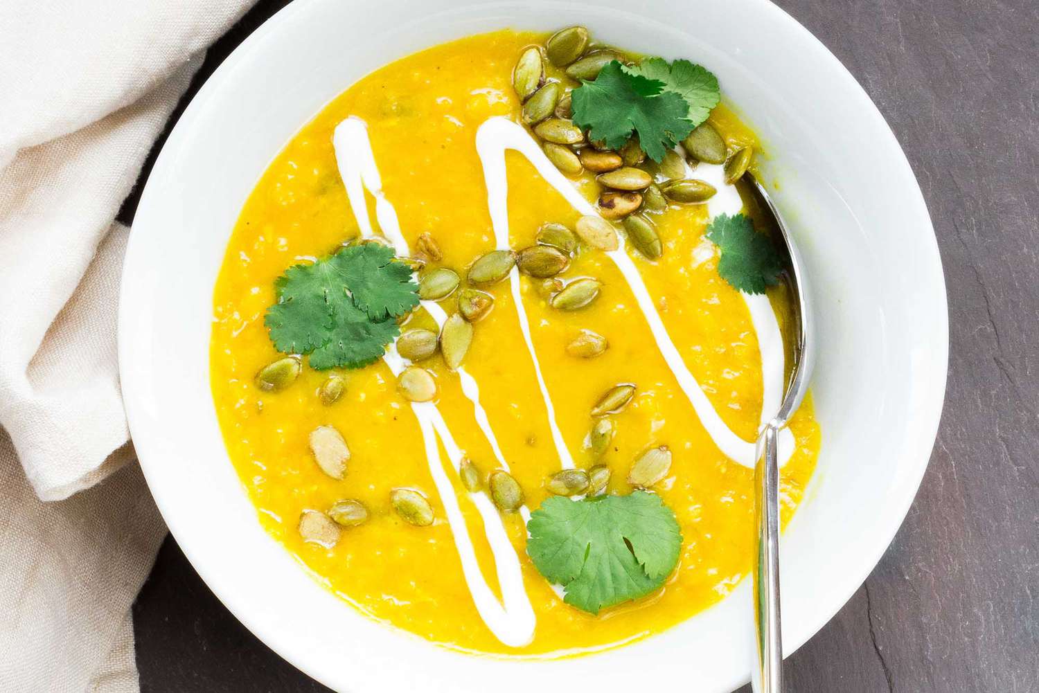 Vibrant yellow split pea soup in a white bowl with cilantro, pumpkin seeds and a drizzle of yogurt