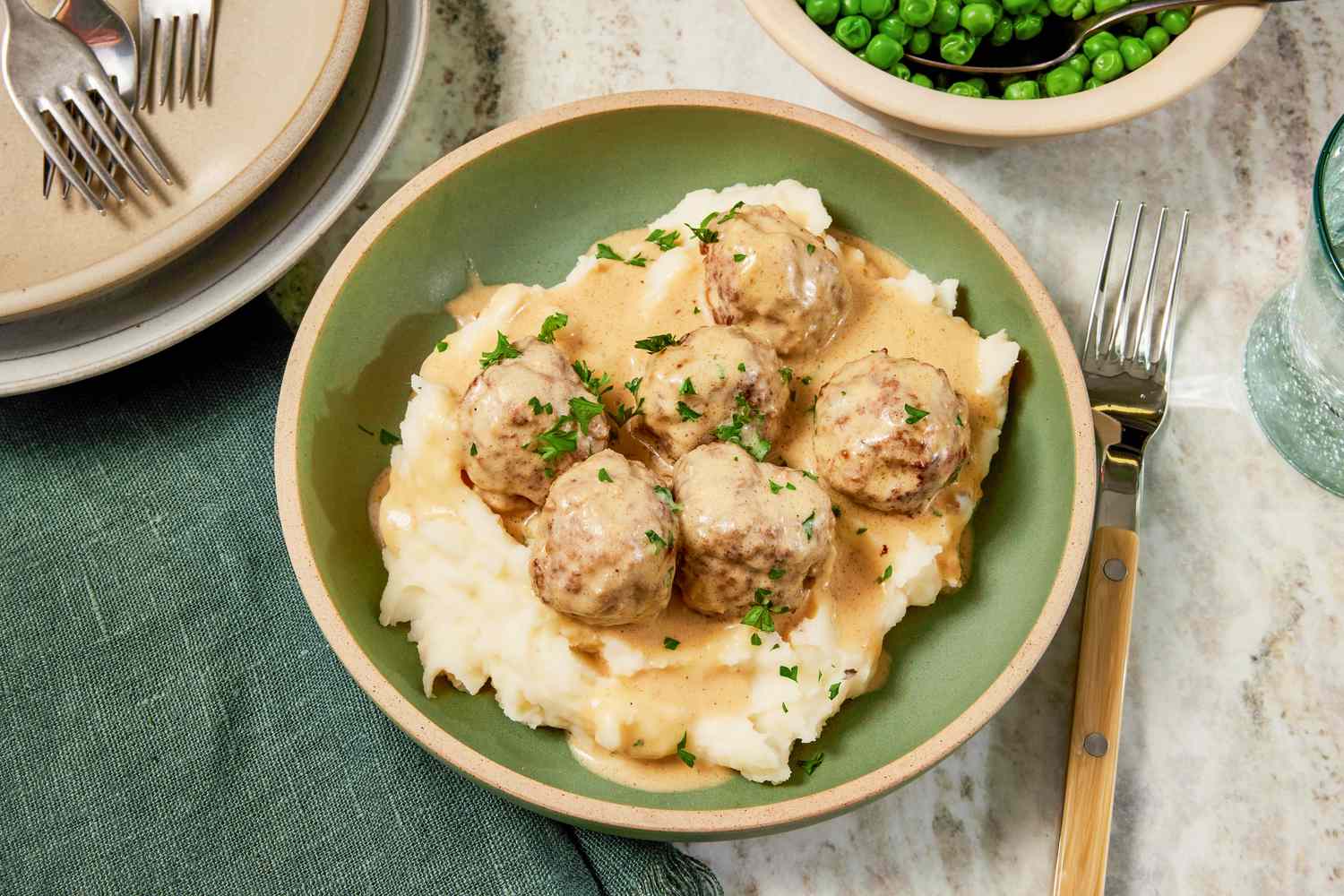 Bowl of copycat Ikea Swedish meatballs served over mashed potatoes, and in the surroundings, a table setting with a bowl of peas, a stack of plates with utensils, and a sage table napkin