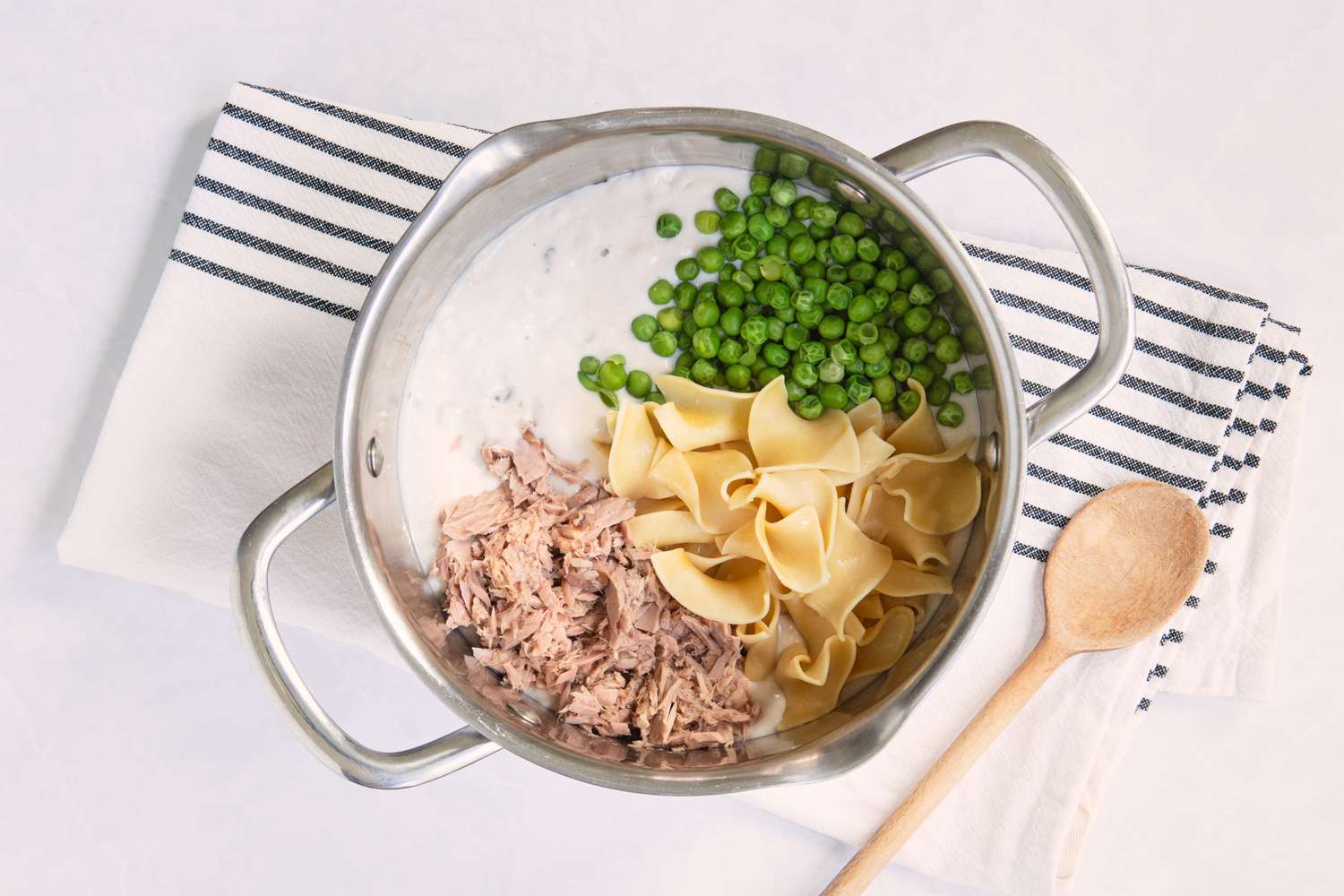 Ingredients for a tuna noodle casserole in a pot including tuna peas and noodles placed on a striped towel with a wooden spoon nearby