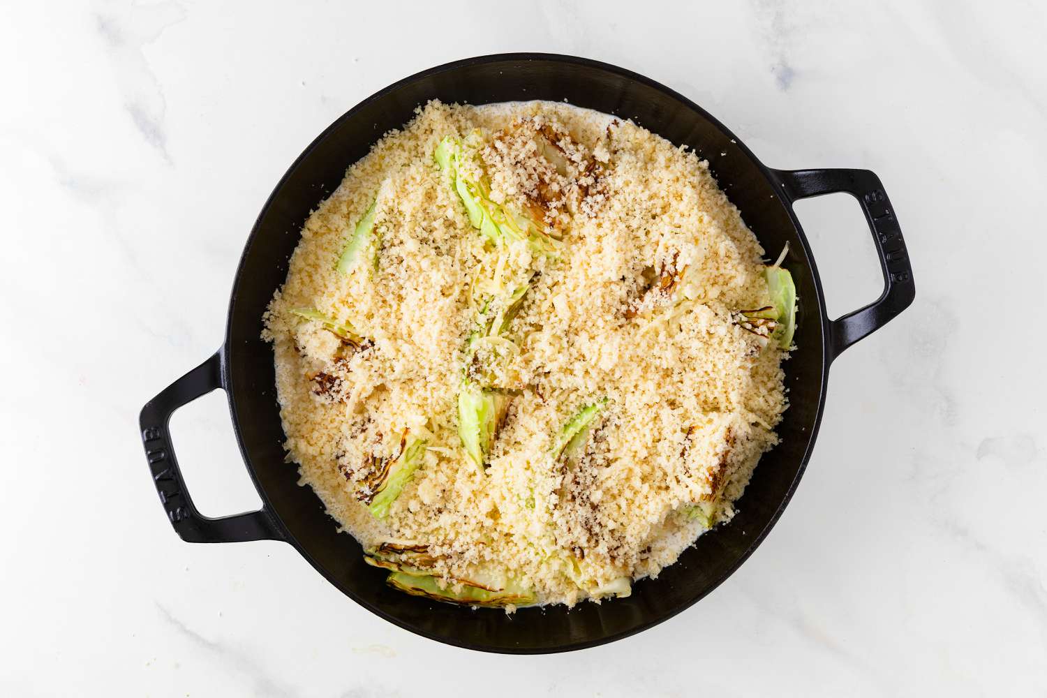 Overhead view of browned cabbage in a cast iron skillet preparing to bake by topping with Parmesan and panko breadcrumb mixture for Cabbage Au Gratin recipe