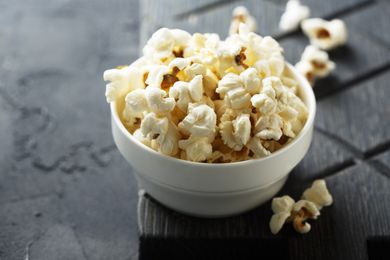 Bowl of popcorn with pieces scattered nearby on a dark textured surface