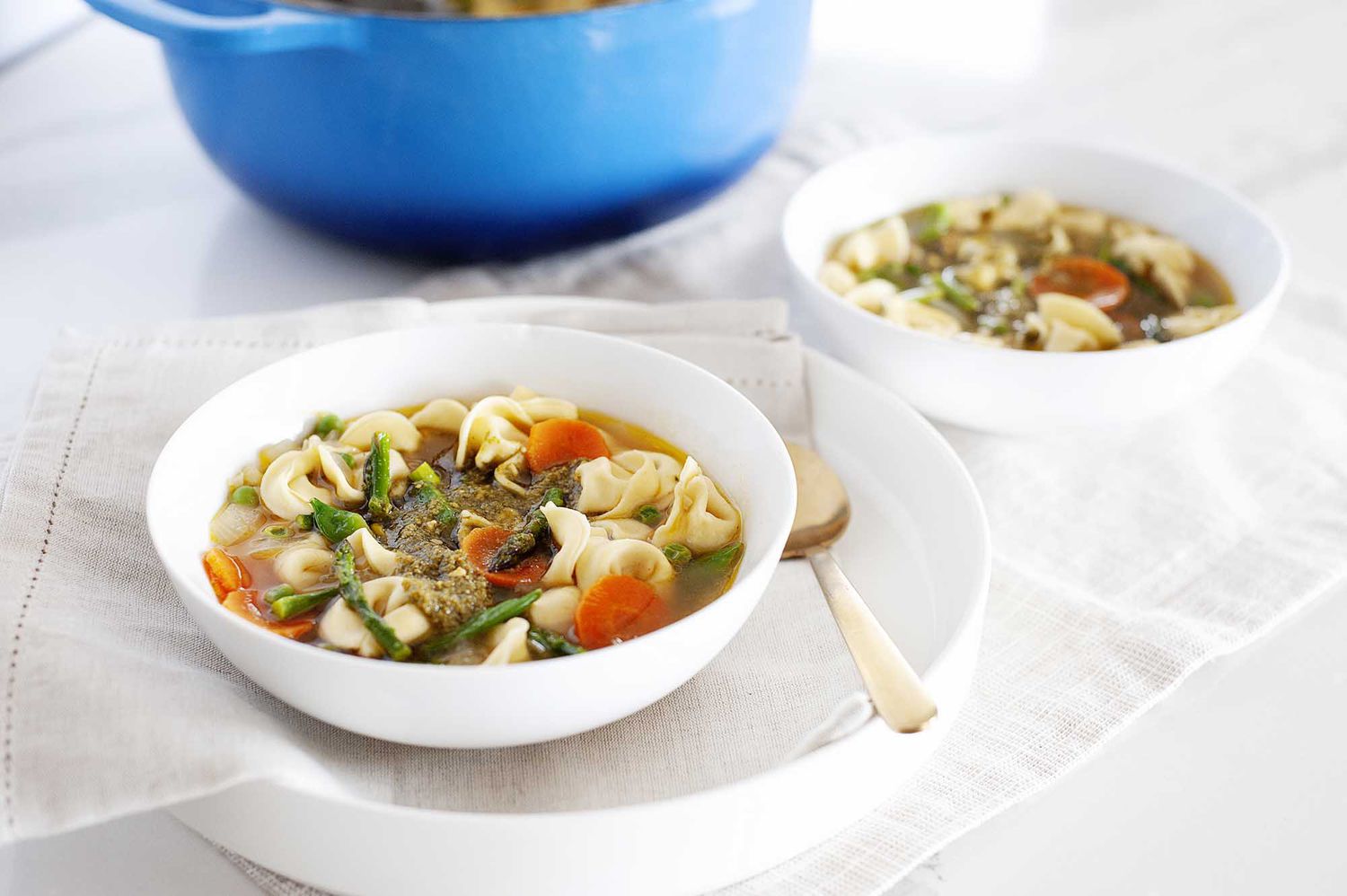 Pesto and tortellini soup with vegetables. A second bowl is to the right and a blue dutch oven is above.