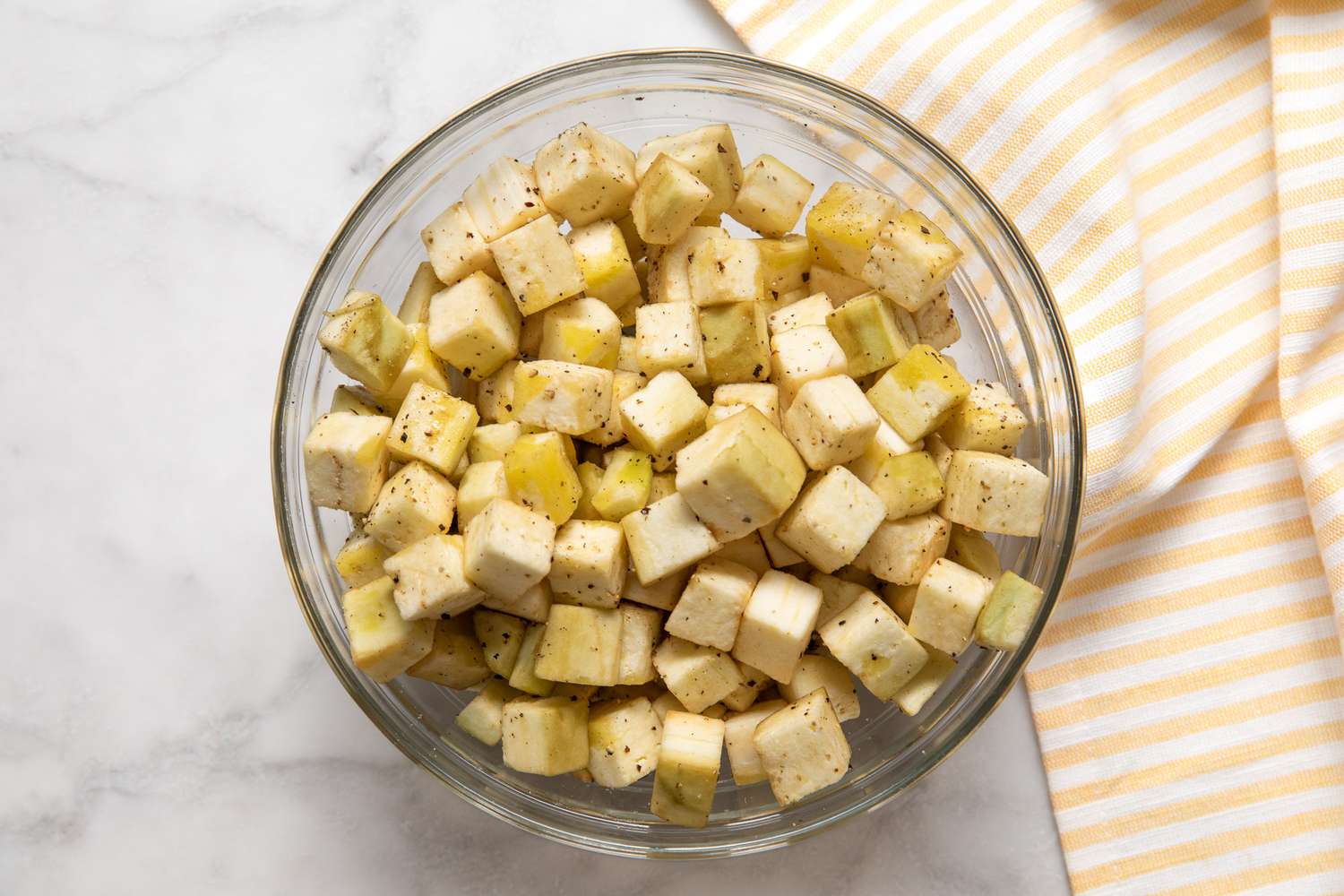 Bowl of Seasoned Cubed Eggplant on a Counter Next to a Yellow and White Striped Kitchen Towel 