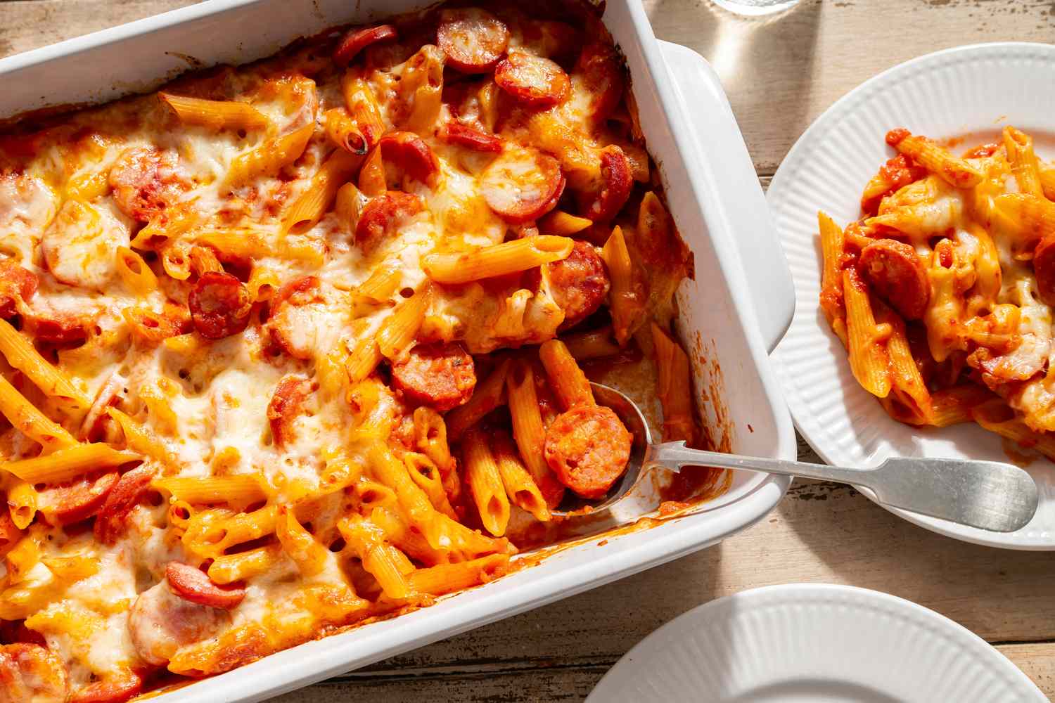 Penne and Smoked Sausage Casserole in a baking dish with a serving spoon, one serving on a dish