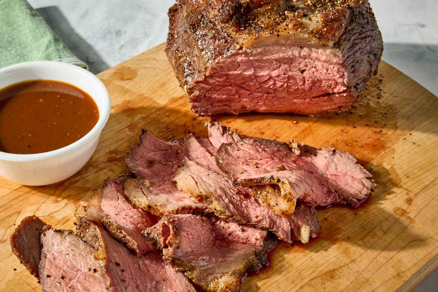 Sliced roast beef on a wooden cutting board with a small bowl of sauce nearby