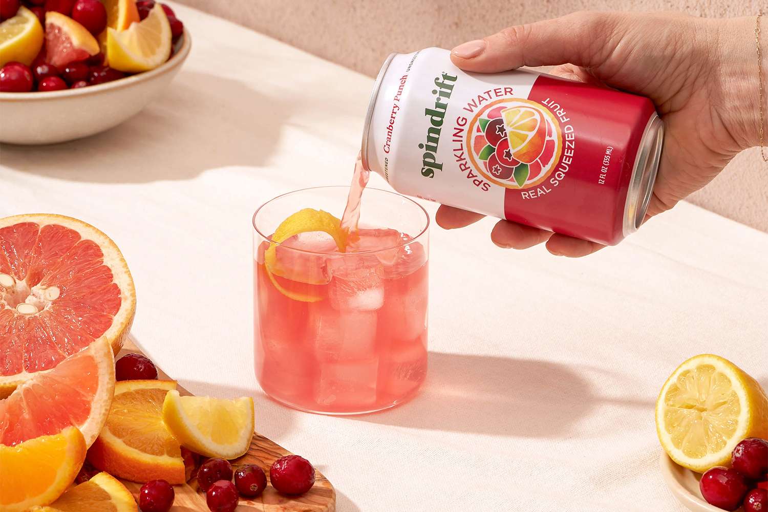 A hand pouring sparkling water from a Spindrift can into a glass with ice surrounded by sliced fruits and cranberries