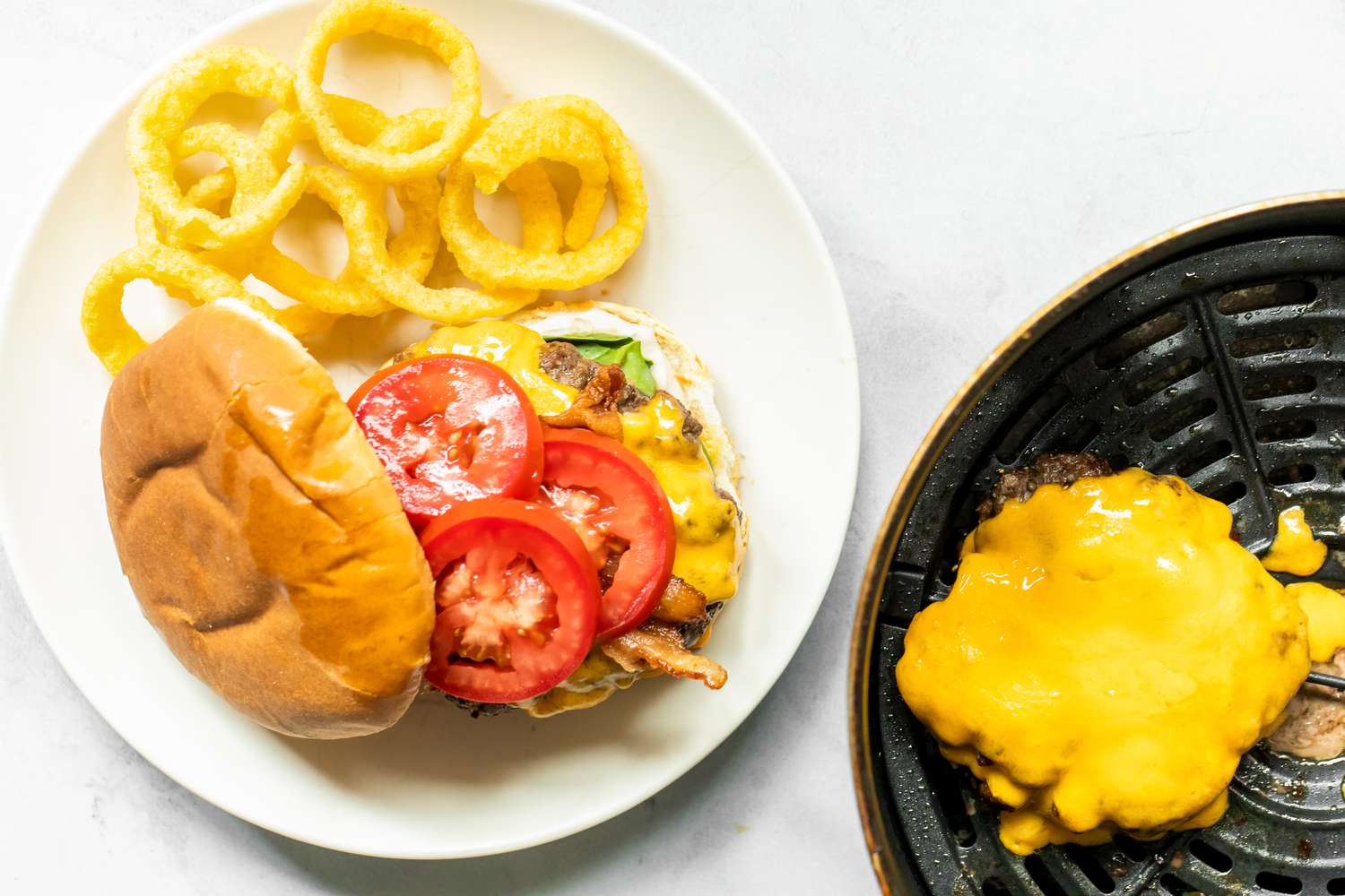 Air Fryer Burger with Onion Rings on a Plate Next to Air Fryer Basin with More Burger Patties