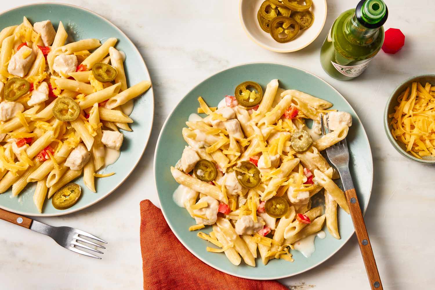 Overhead view of rattlesnake pasta topped cheese and jalapenos in two light green plates along with a fork and next to an orange napkin, bowl of jalapenos, bowl of cheese and a bottle of hot sauce on a beige background