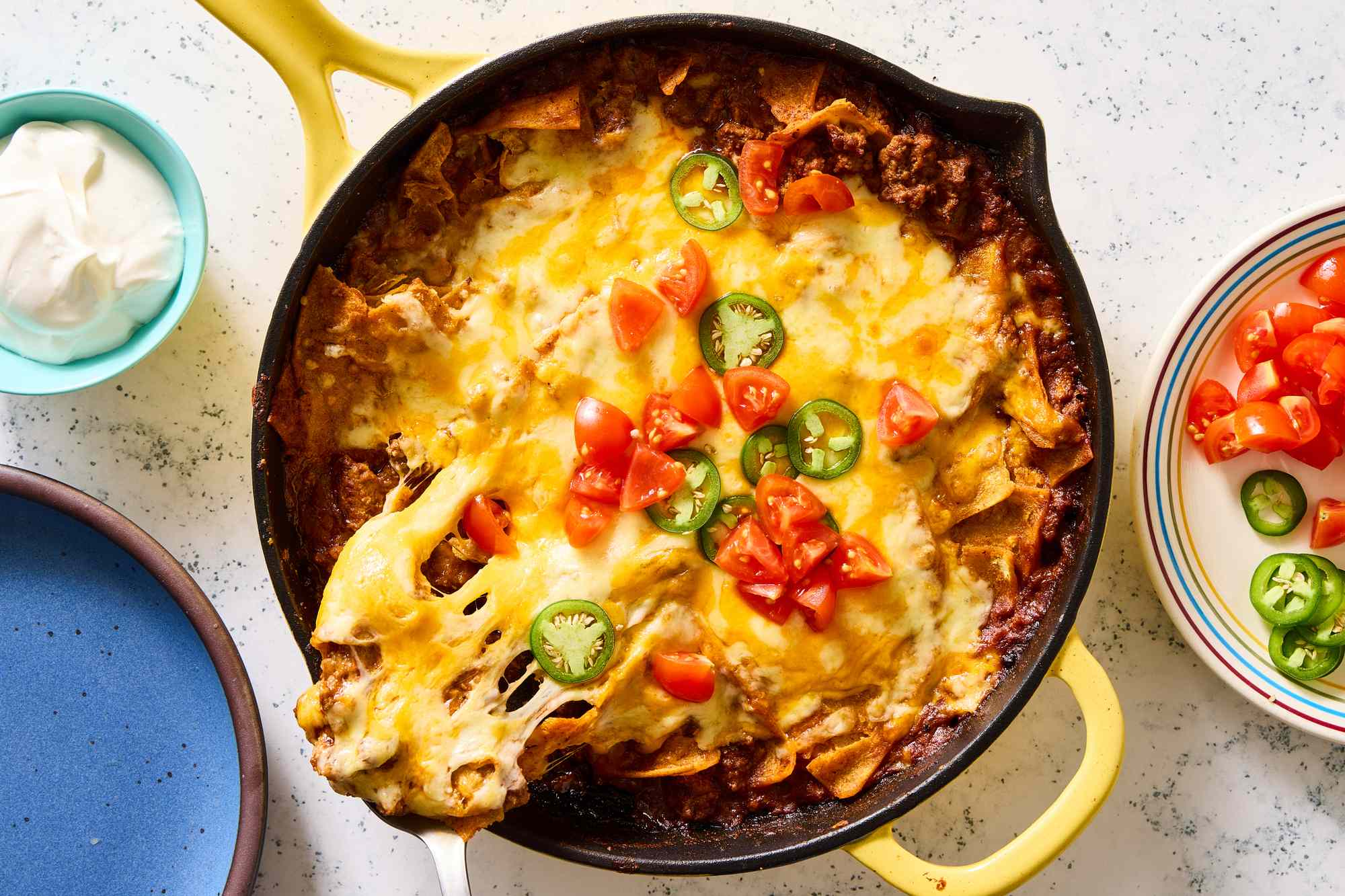 A skillet dish topped with melted cheese, tomatoes, and jalapeños, placed alongside small bowls of toppings