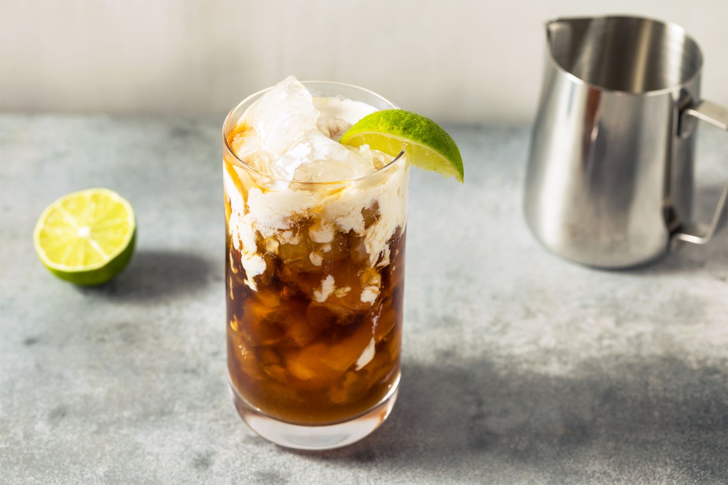 A glass filled with a dark beverage ice and lime wedge with a lime and a metal pitcher in the background