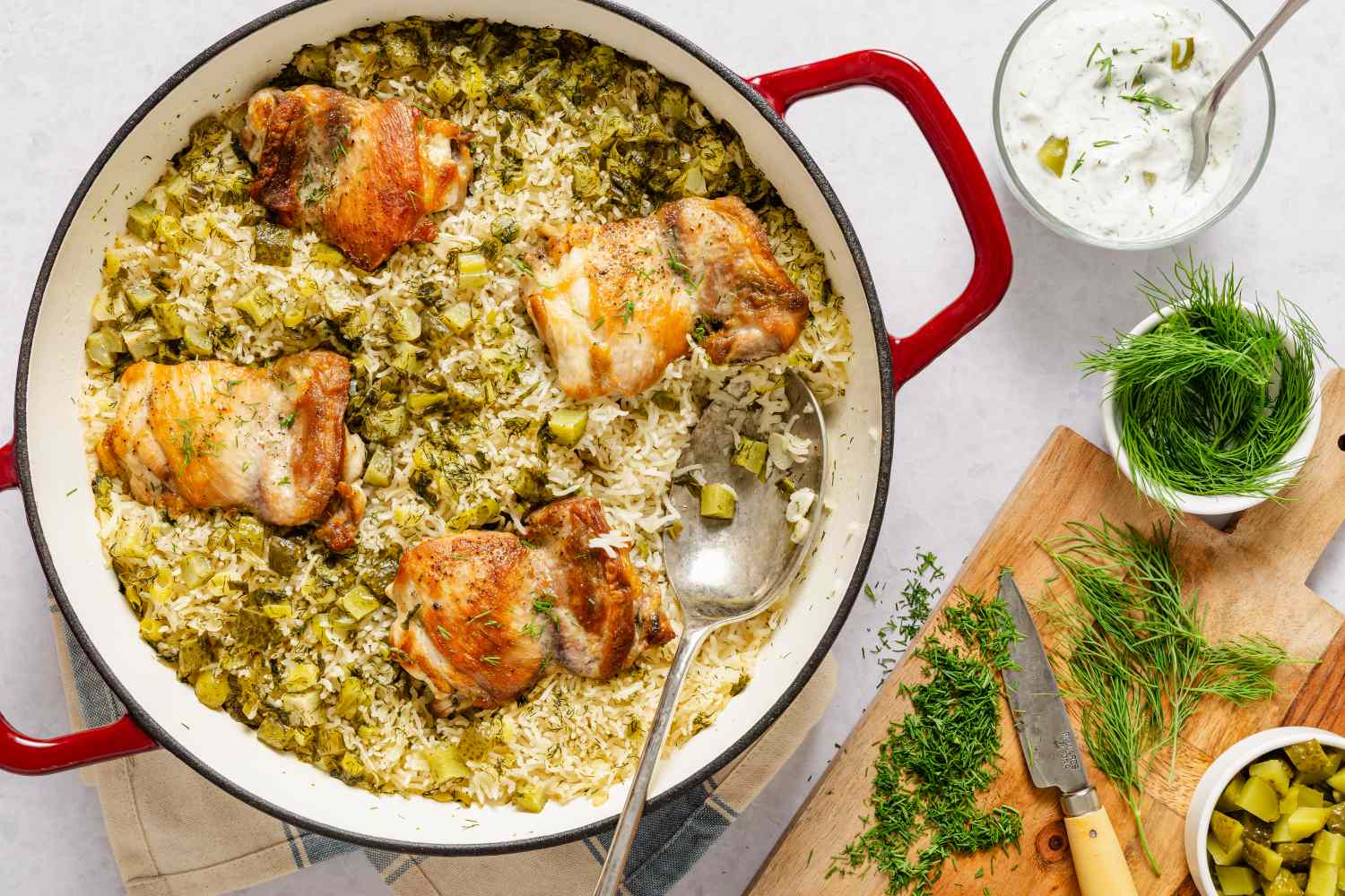 A skillet with dill pickle chicken and rice garnished with fresh herbs accompanied by a cutting board with dill and pickles