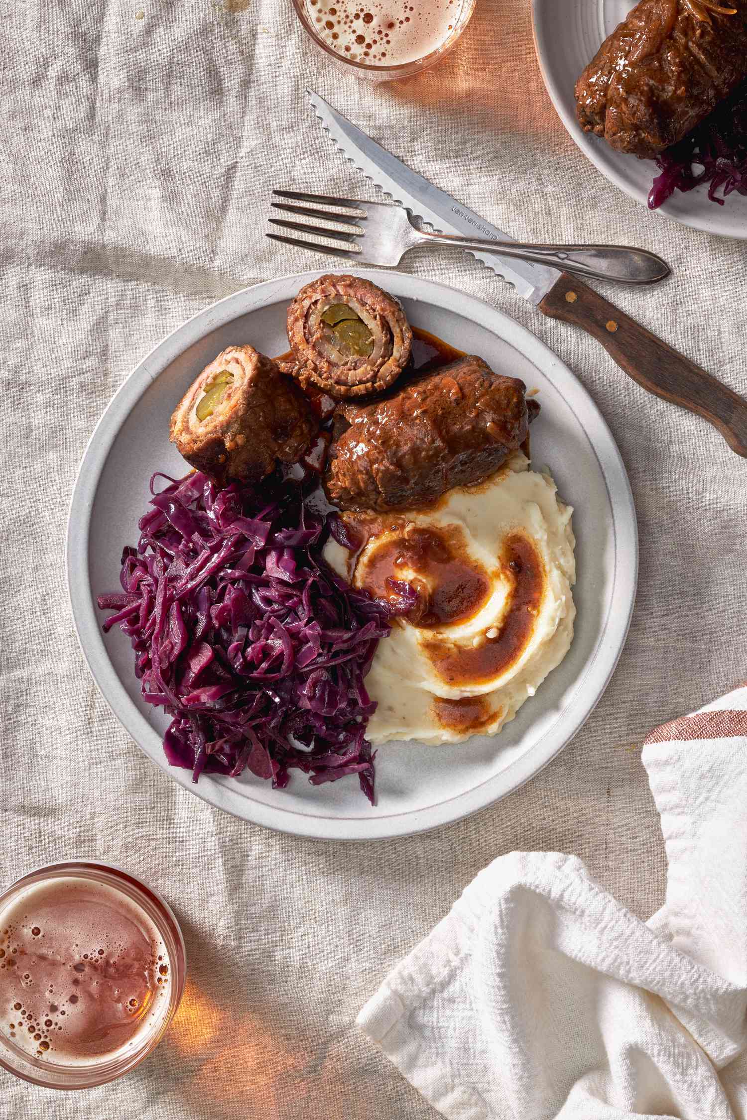 German rouladen with cabbage and mashed potatoes and gravy on a plate at a table setting with another serving on a plate, a glass with beer, and utensils
