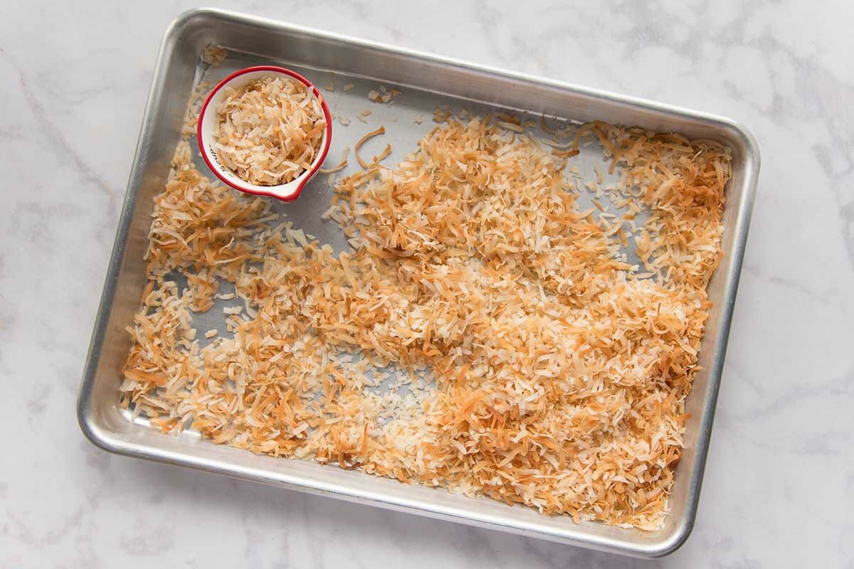 Shredded coconut is toasted on a baking sheet. A small cup has additional coconut inside on the sheet pan.