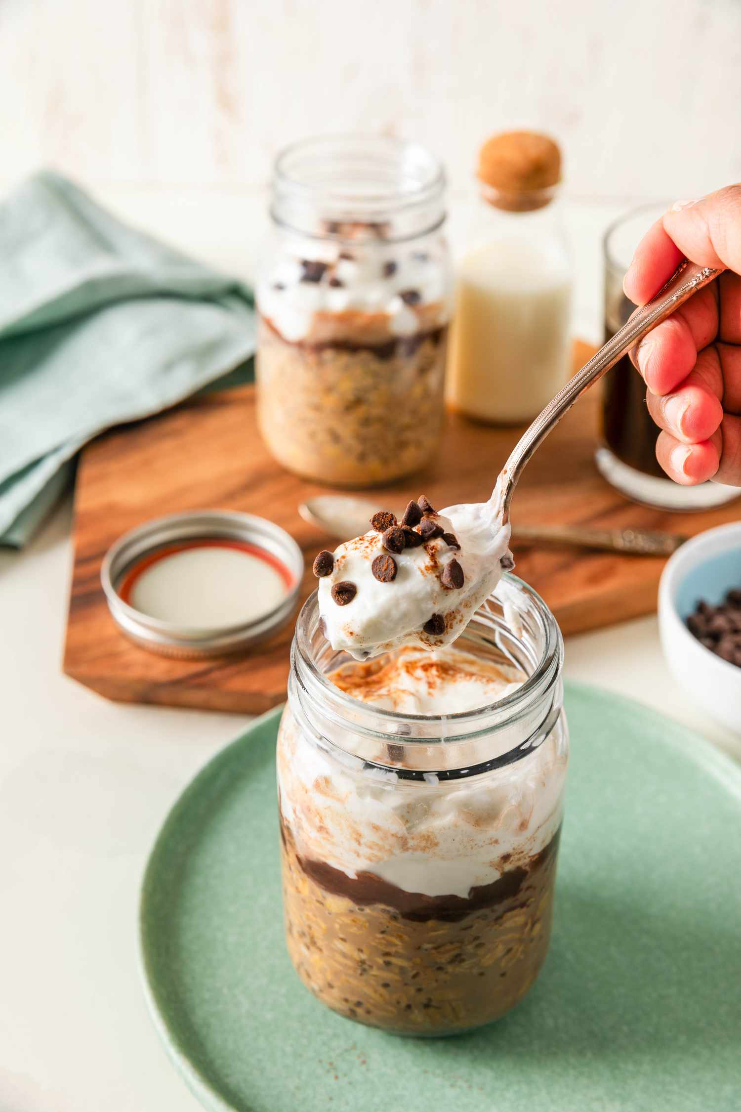 A spoonful of tiramisu overnight oats lifted from the jar on a small plate, and in the surroundings, on a wooden board, another jar of overnight oats, a small carafe of milk, a glass of coffee, and a robin blue kitchen towel
