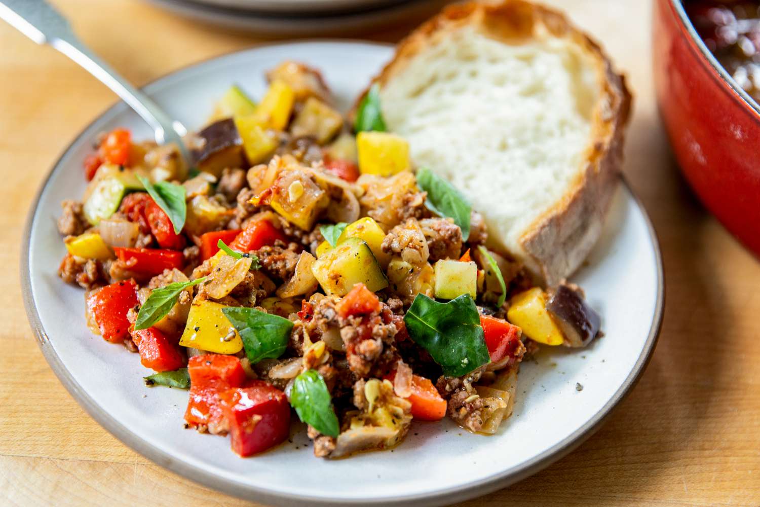 A cooked dish on a plate featuring diced vegetables, ground beef, and a slice of bread with a fork to the side