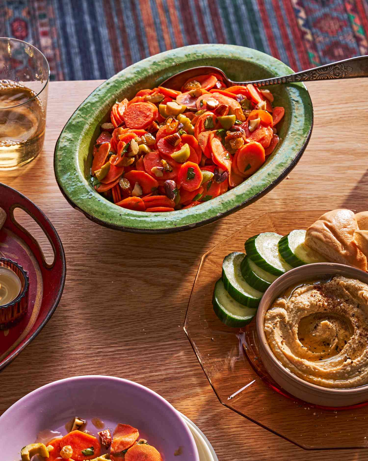 A bowl of carrot salad on a coffee table, alongside hummus with cucumbers and pita and a glass of wine