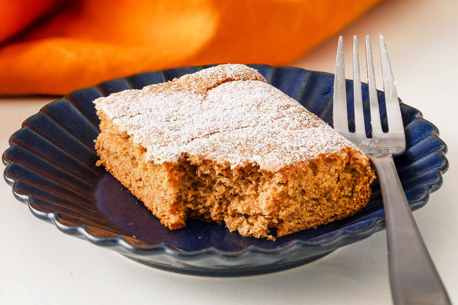 A slice of soda cake topped with powdered sugar on a blue plate accompanied by a fork