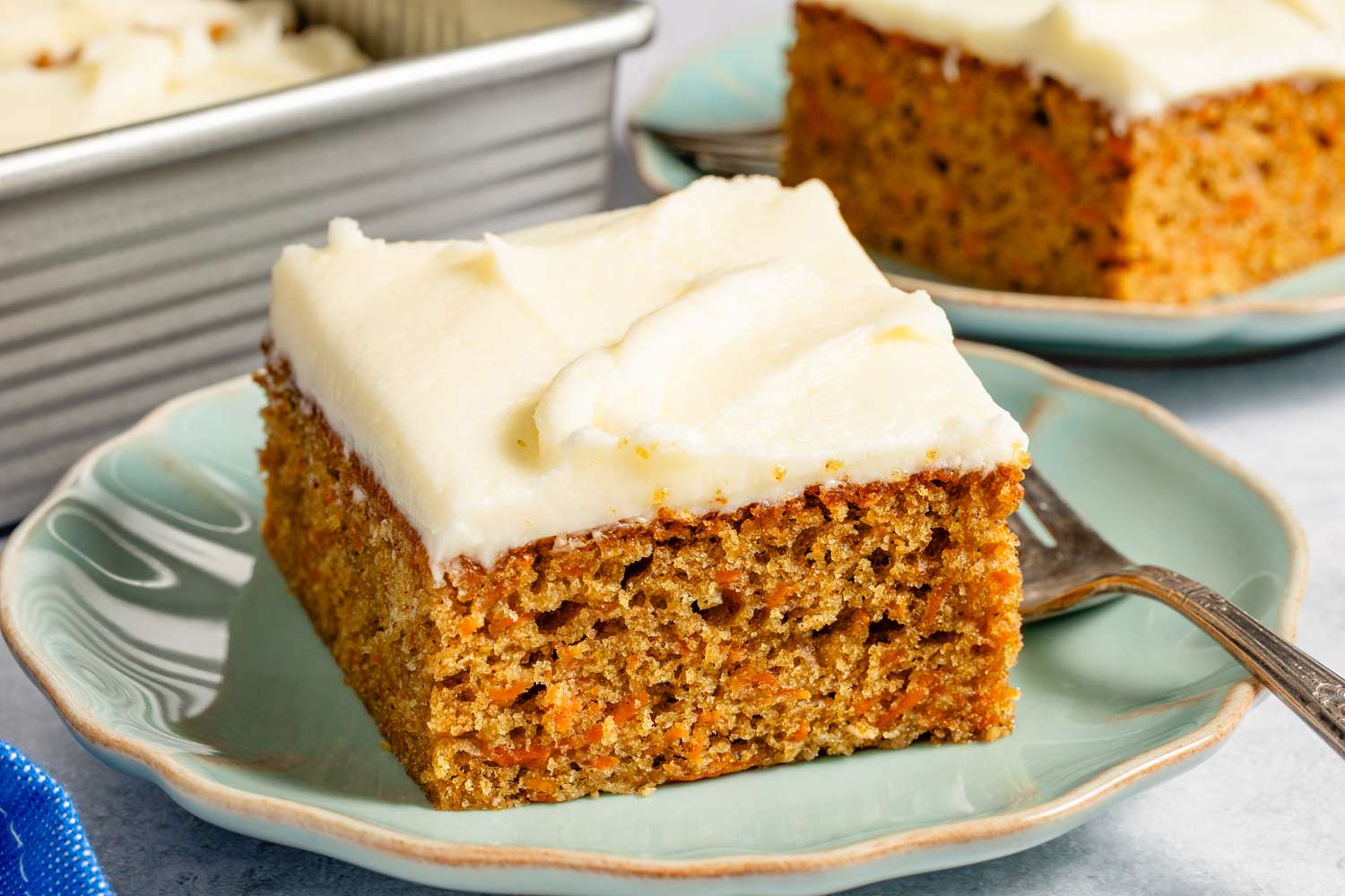 Carrot cake slice with cream cheese frosting on a plate, fork beside it, baking dish in the background.