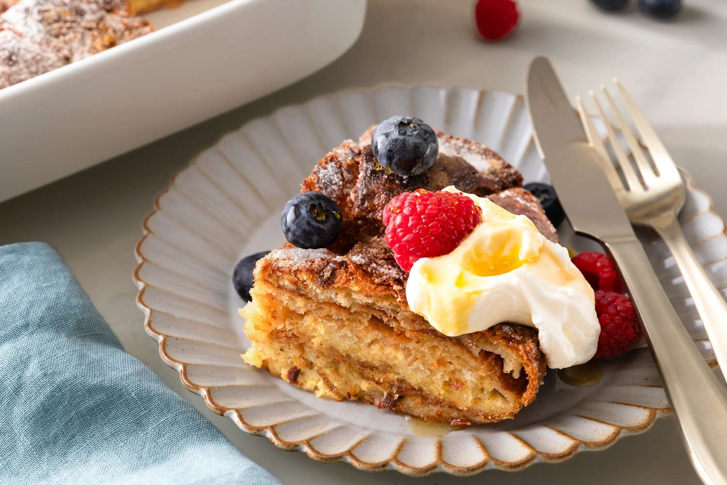 Closeup view of a small plate with a slice of Croissant French Toast casserole topped with fruit and butter next to a knife and fork