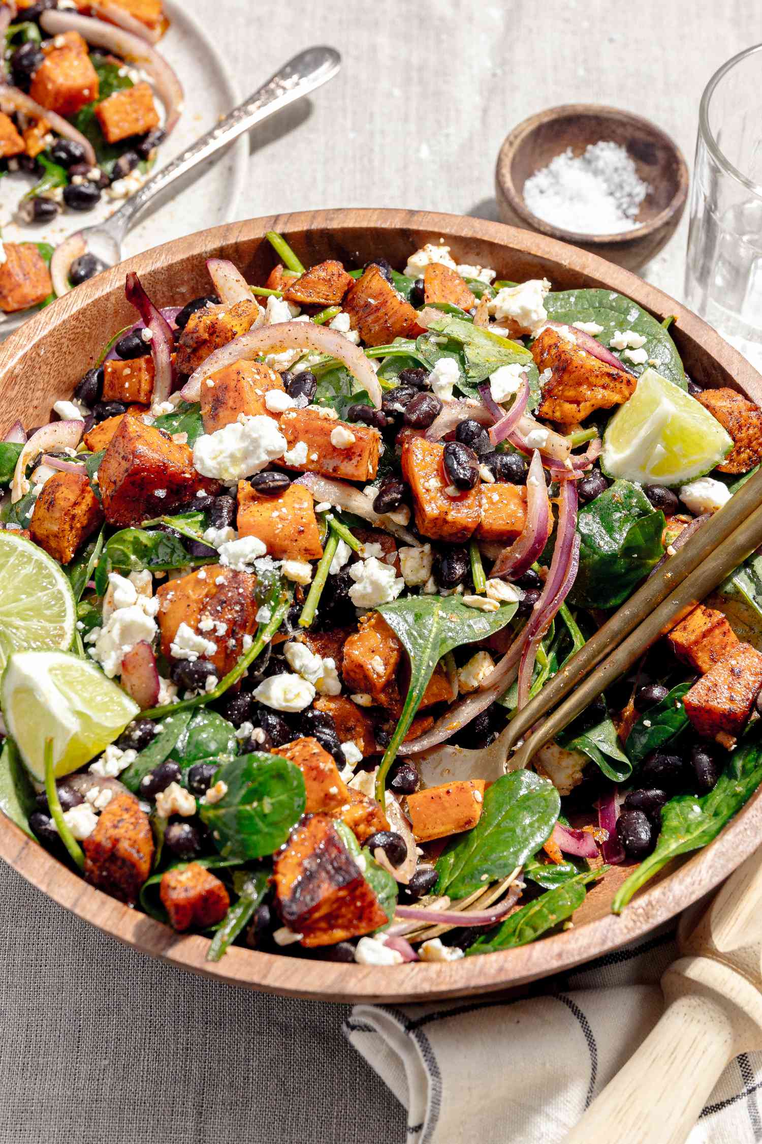 Bowl of Roasted Sweet Potato Salad Topped with Crumbled Feta Cheese and Lime Wedges, Surrounded by a Plate with Some Salad, a Small Bowl of Salt, and a Kitchen Towel 