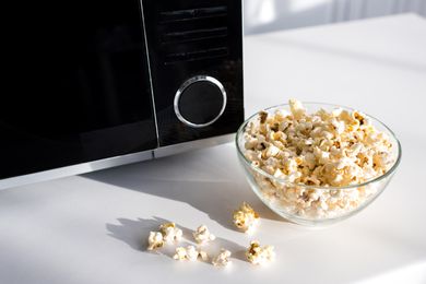 A bowl of popcorn on a counter next to a microwave some popcorn scattered outside the bowl