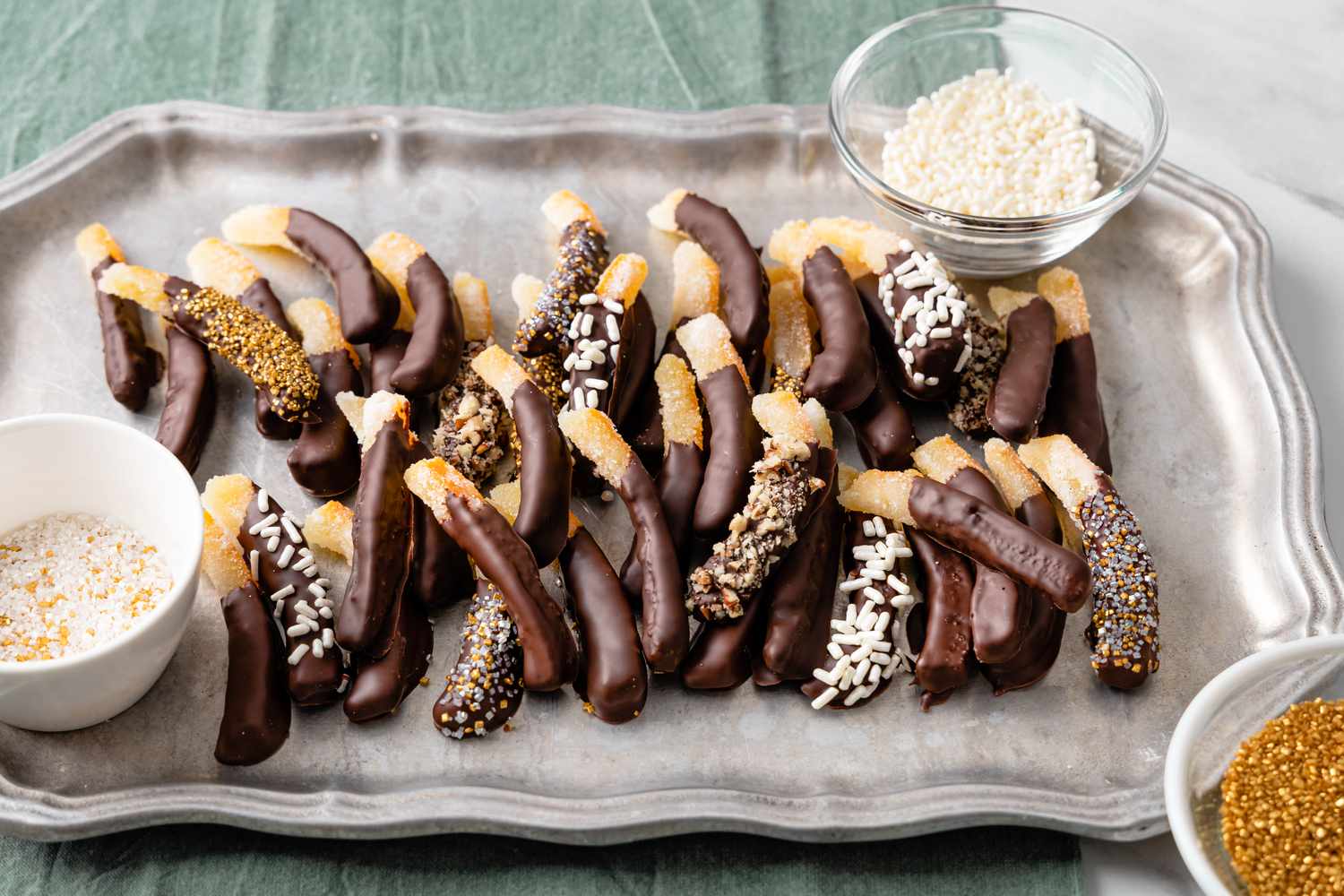 Chocolate Covered Orange Peels on a Tray with Two Bowls of Toppings. Some Chocolate Covered Orange Peels Are Topped with Sprinkles and Nuts.