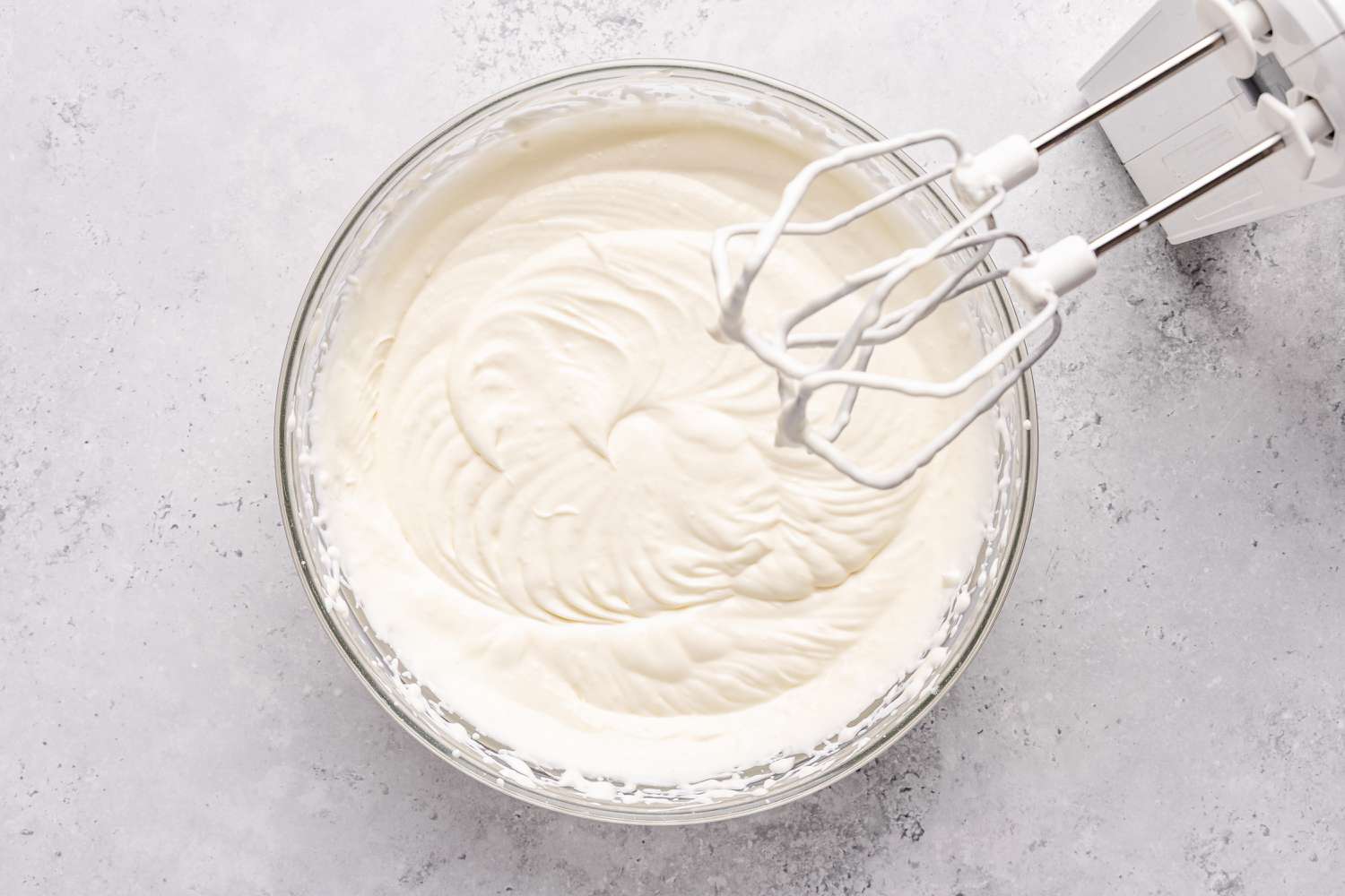 Overhead shot of whipped cream in a clear mixing bowl with a electric wisp next to it for the stained glass jello recipe