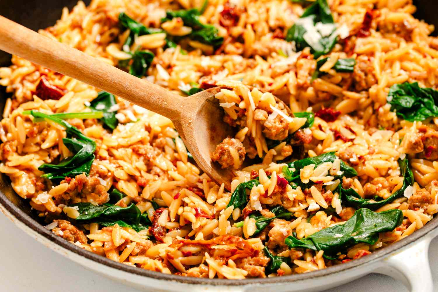 Angled view of a wooden spoon in a skillet of orzo, italian sausage, sun-dried tomatoes and baby spinach