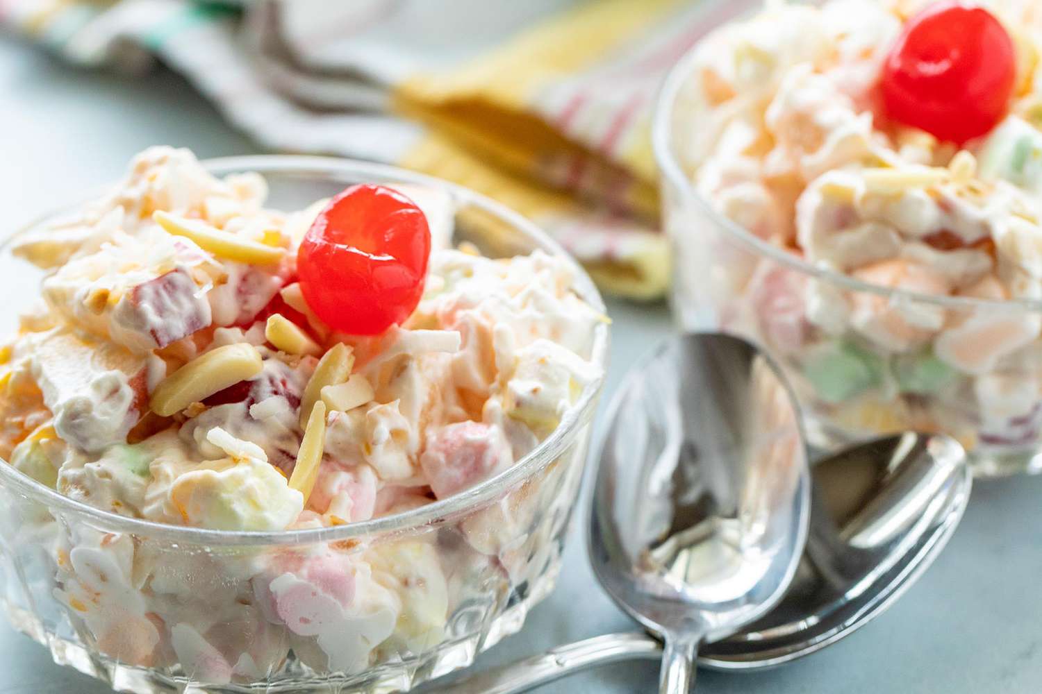 Two small bowls of marshmallow salad with cherries on top are at the front of the image. Two spoons are crossed to the right of the first bowl. A striped dish towel is in the background.