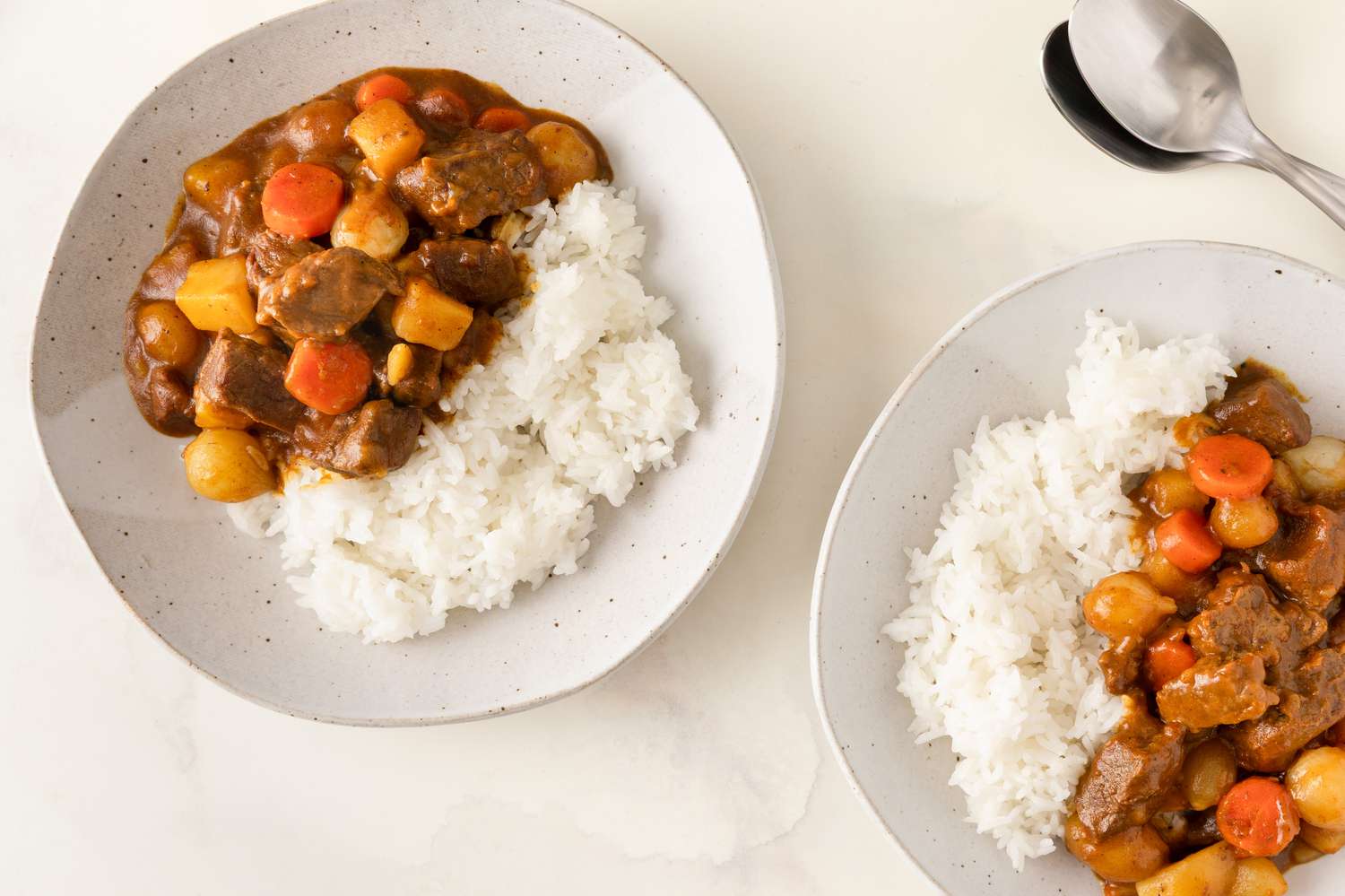 Two plates of rice served with Korean curry having carrots, potatoes, and meat, accompanied by a spoon and fork on the side