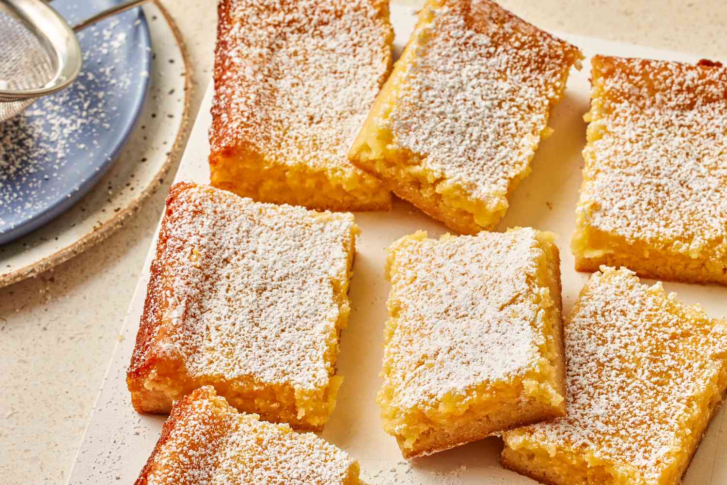 lemon gooey butter cake slices on a platter next to a stack of plates and a colander 