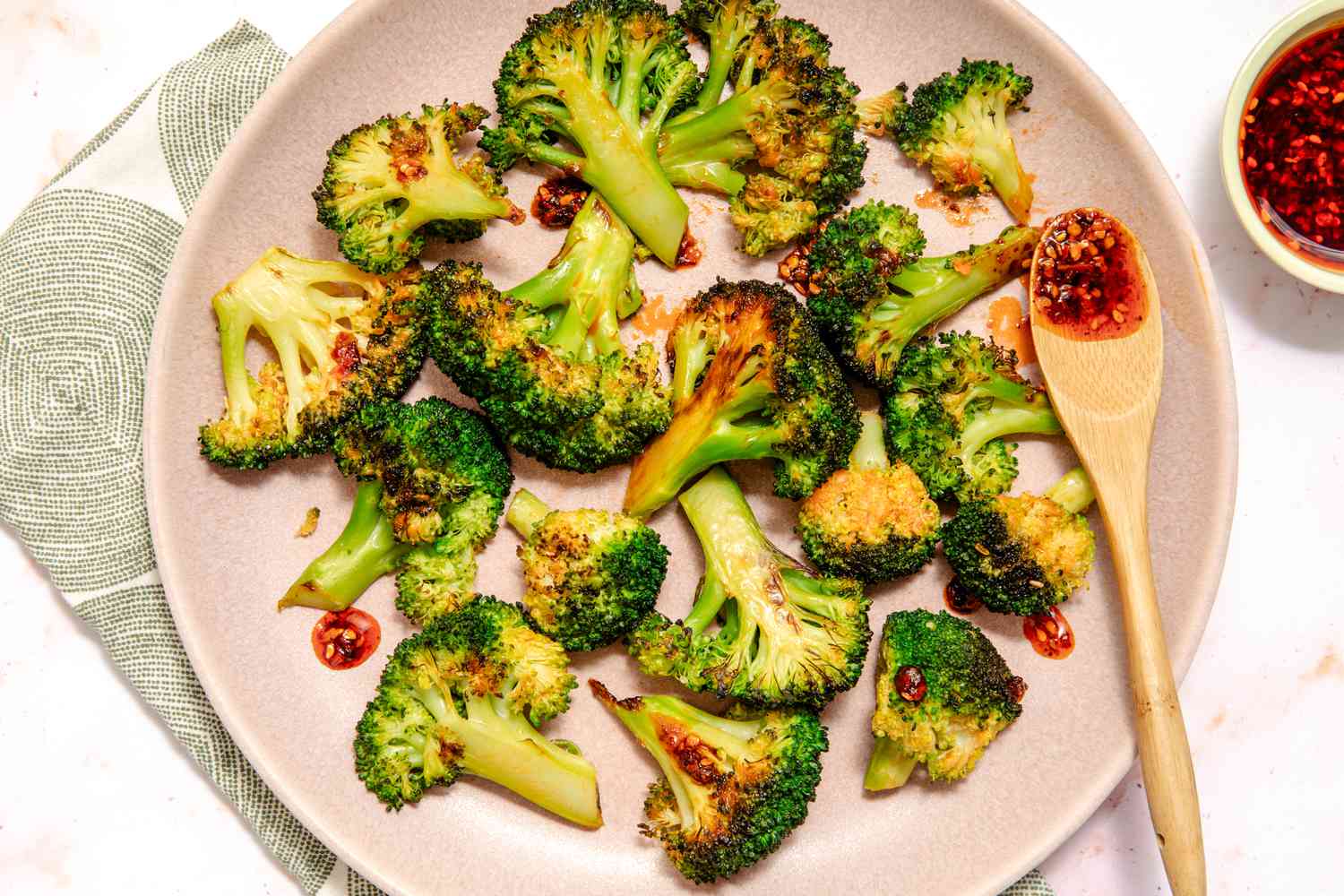 Overhead view of a pink plate of chili crisp coated broccoli florets and a small wooden spoon with chili crisp all resting on a cloth napkin and marble countertop