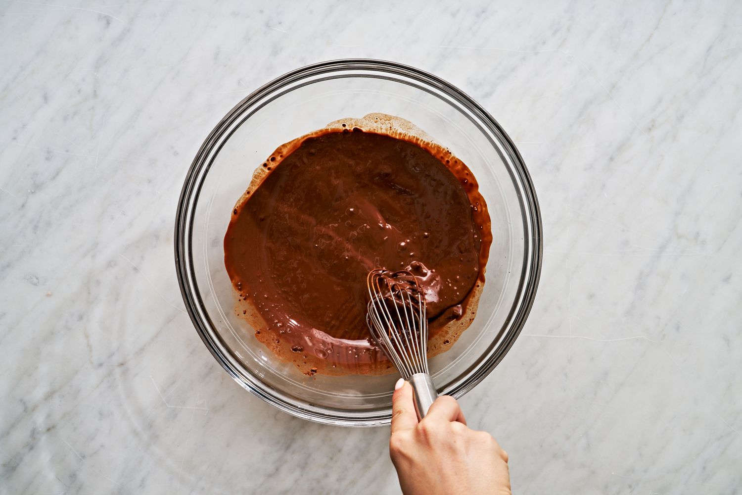 Overhead view of a hand whisking hot coffee and cocoa powder together in a clear glass bowl for Mississippi Mud Cake recipe
