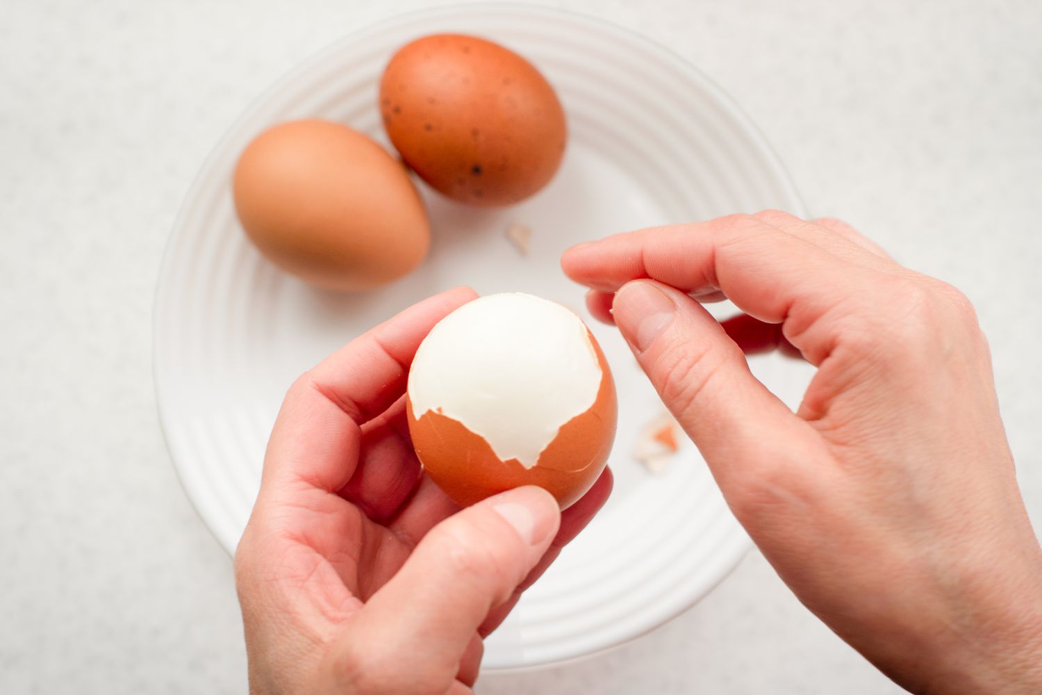 Hands peeling a boiled egg, plate with eggs in the background
