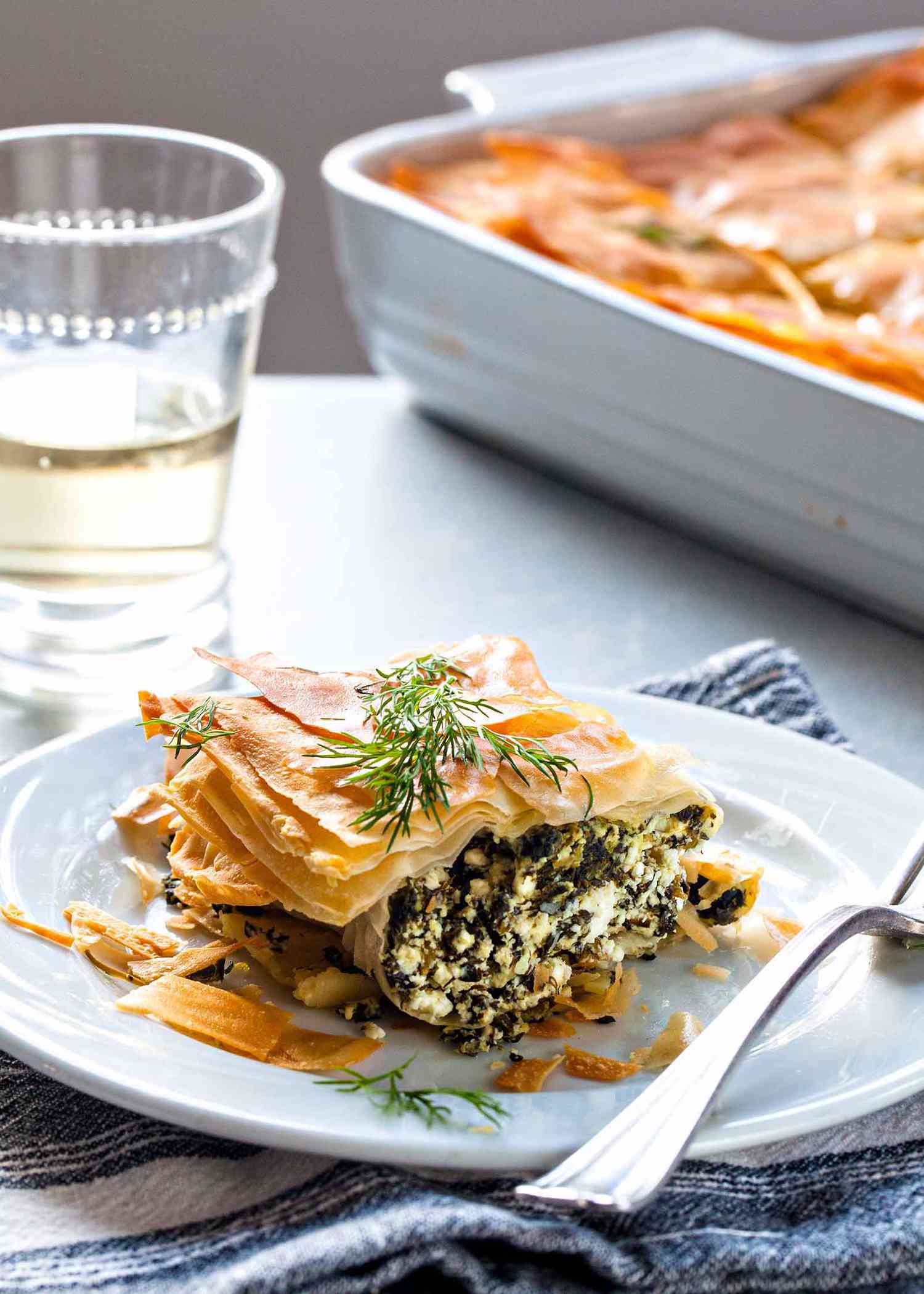 Slice of Greek Spinach Pie on a white plate, with a fork, a glass of white wine, and the casserole dish in the background,