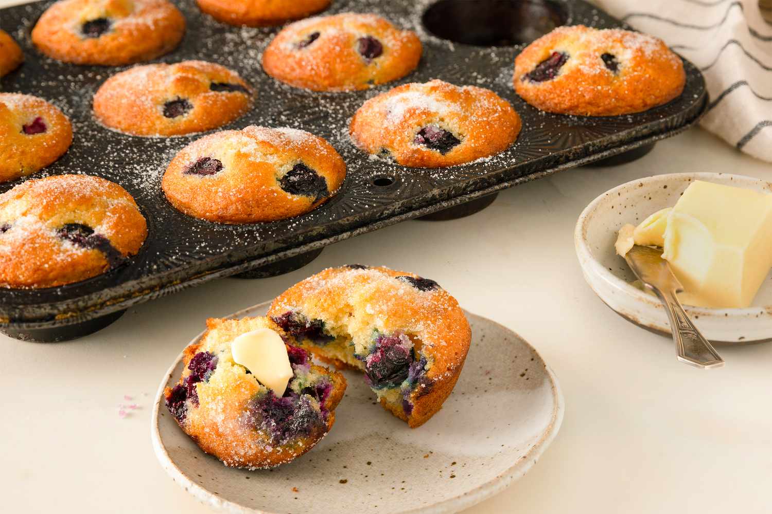 blueberry muffin, torn open and buttered, on a small plate, muffin tin with muffins in the background