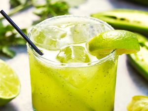 Glass of a green margarita with garnish, surrounded by lime slices and herbs