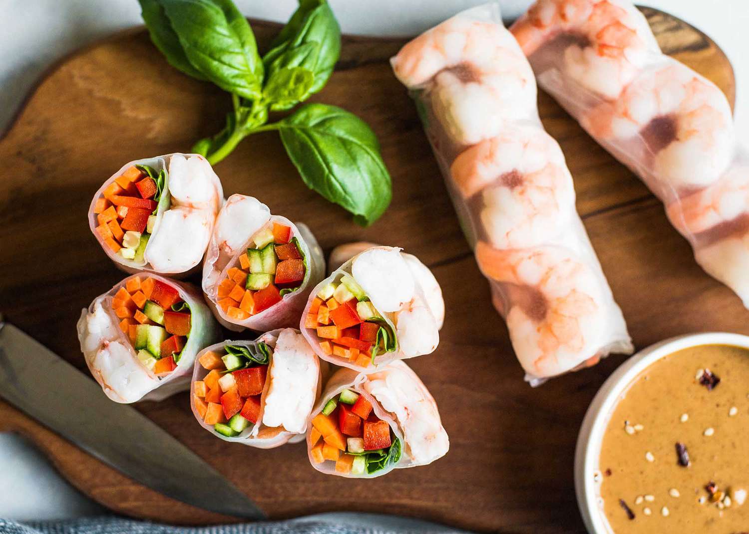 Shrimp Spring Rolls (Some Cut Into Pieces With Cross Section Facing Up and Some Still Whole) on a Wooden Board With a Bowl of Peanut Sauce and a Sprig of Basil