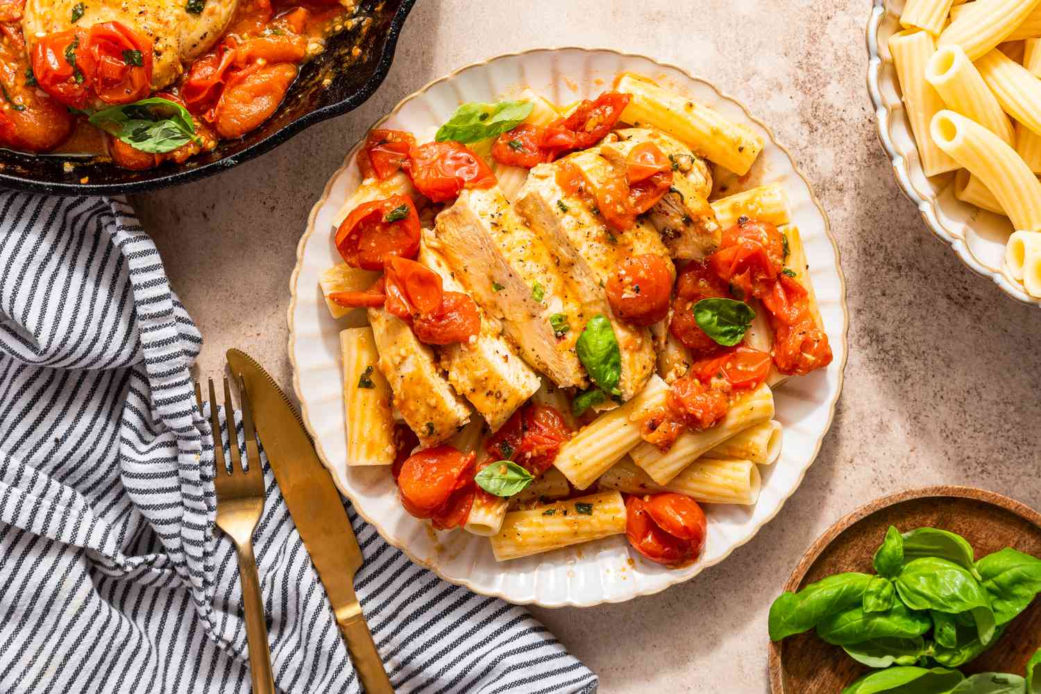 chicken pomodoro over rigatoni on a plate at a table setting with more in a cast iron skillet, a bowl of rigatoni, bowl of basil, and a table napkin