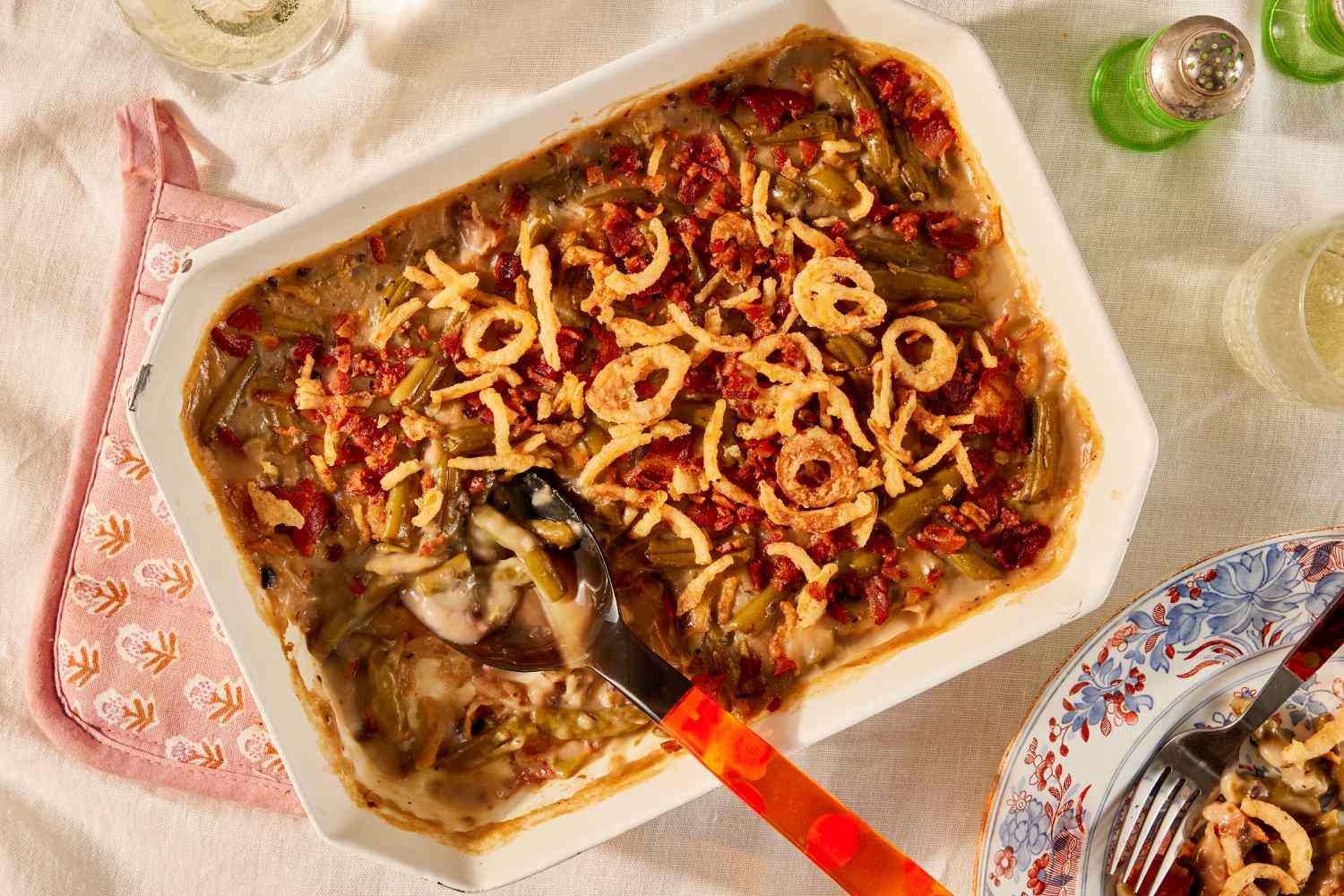 Overhead view of a beige casserole dish with green bean casserole along with a red-handled serving spoon resting on a pink hot pad and beige table cloth