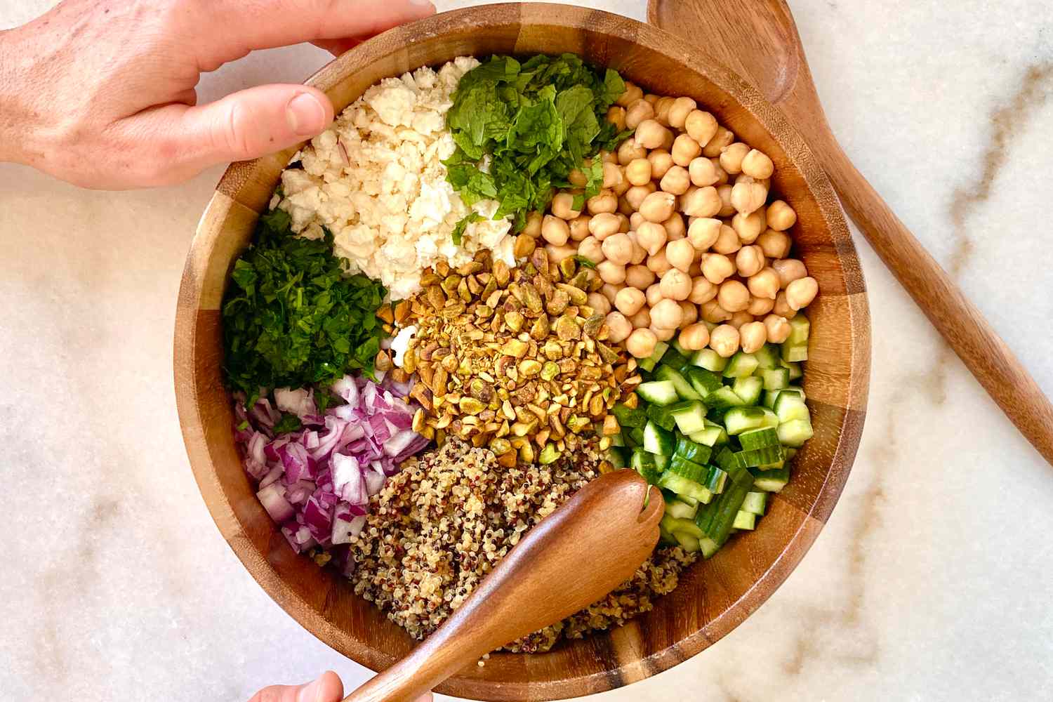 A wooden bowl filled with piles of salad ingredients: chickpeas, cucumber, quinoa, red onion, parsley, feta, mint, and pistachios