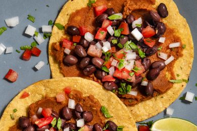 Two tostadas topped with refried beans black beans diced tomatoes onions and herbs on a plate with a lime wedge nearby
