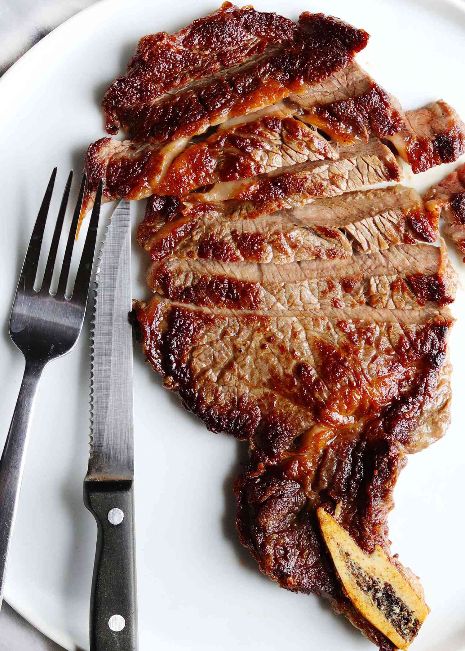 Bone in steak cooked on a stovetop and set on a white plate. A steak knife and fork are to the left of the sliced steak. The steak is cooked to medium and has a dark crust.
