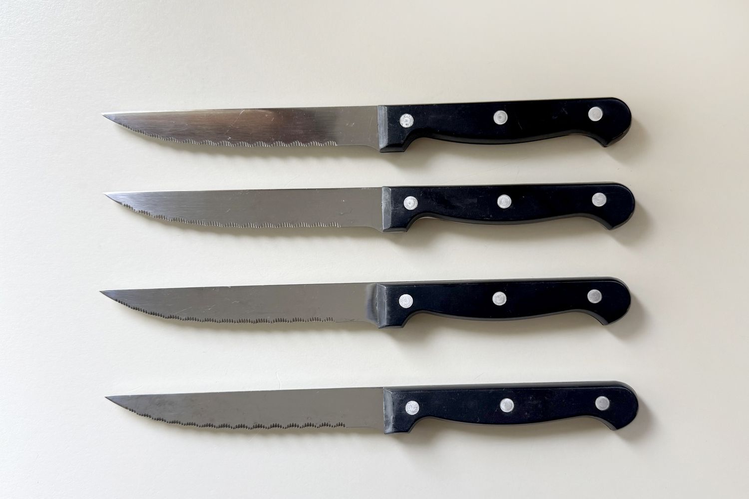Four steak knives with serrated edges and black handles arranged in a row