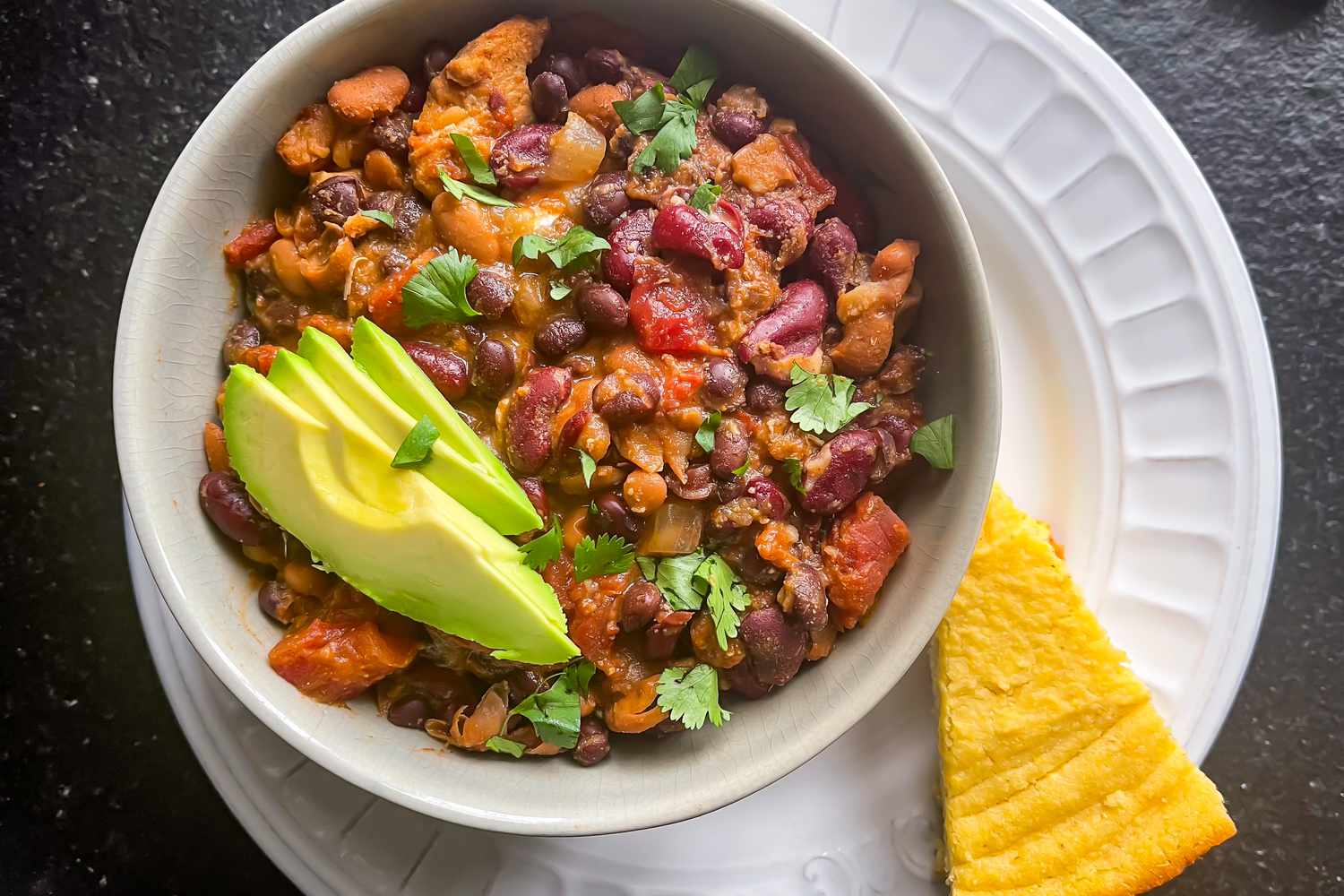 A bowl of chili with beans, avocado slices, and herbs, served with a piece of cornbread