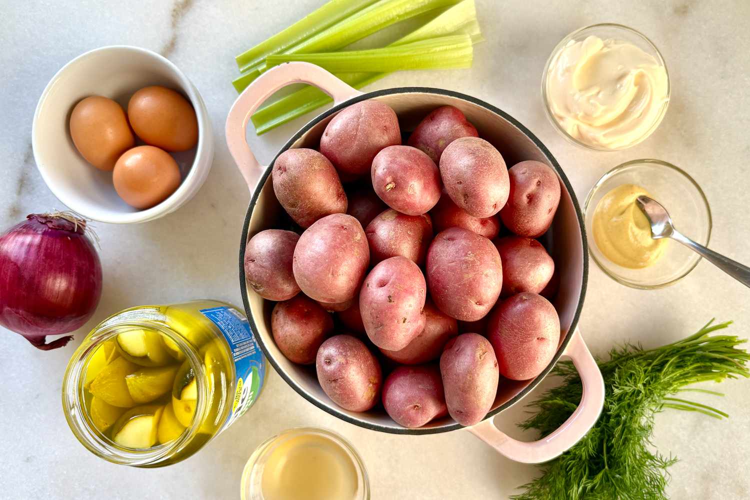 Ingredients for a dish include potatoes, eggs, celery, red onion, dill, pickles, mayonnaise, and mustard arranged on a surface