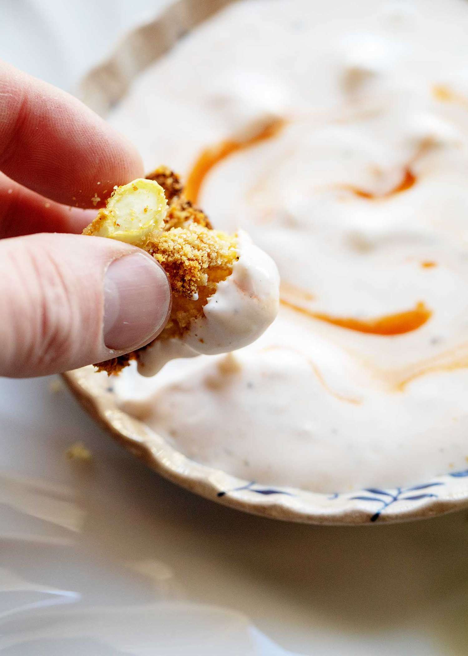 Thumb and forefinger holder air fried cauliflower that has been slightly dipped in a creamy sauce. The sauce is in a fluted bowl and swirled with hot sauce.