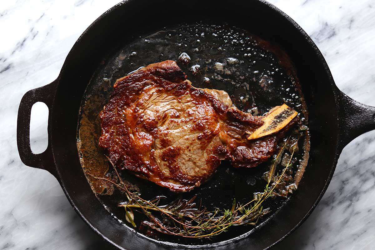A cast iron skillet with steak cooked on the stove. Oregano sprigs are at the bottom of the pan. Oil and juices are visible in the pan.